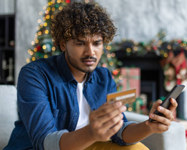 A man is sitting on his couch, holding his phone and his credit card while looking confused.