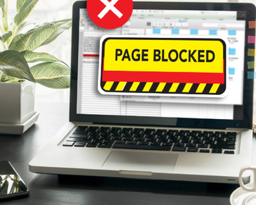 An open laptop on a work desk with the screen displaying the text &quot;page blocked&quot; over a restricted website
