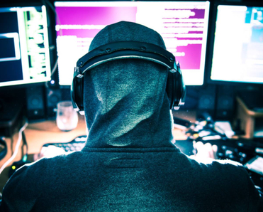 A hooded hacker viewing three computer monitors. 