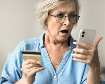 A shocked older woman is holding her smartphone in one hand and her credit card in another.
