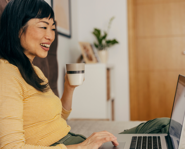 A woman holds a coffee mug and uses her laptop in bed.
