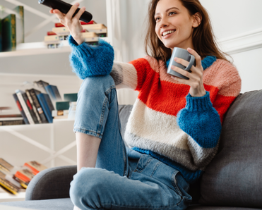 A woman sits on her couch, holding a mug and the remote.