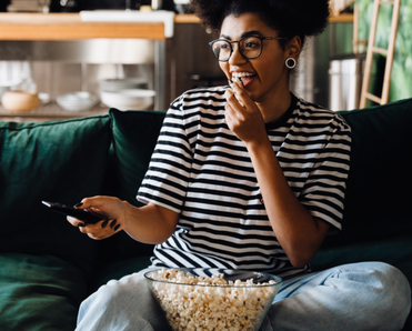 A woman sits on the couch, eating popcorn and watching TV.