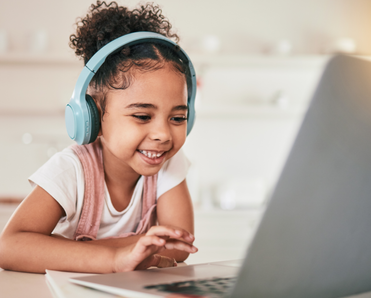A little girl wears headphones while watching playing on her laptop. 