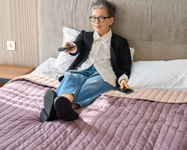 An older woman watches TV in a hotel room.