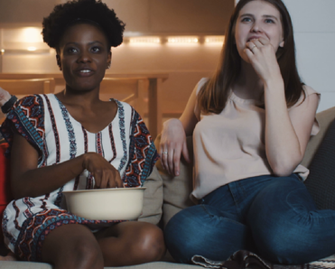 Happy young multiethnic women friends sit together at home watching movies smiling together with snacks slow motion.