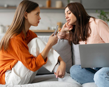 Mother sitting on sofa with laptop spying on teen daughter using cellphone, mom and kid spending leisure time together on devices.