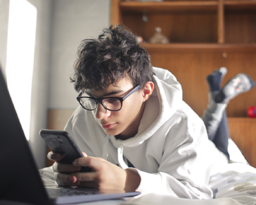 Teen boy laying on bed while scrolling on his smartphone and working on his laptop.