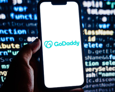 Closeup of smartphone displaying GoDaddy logo with code on a screen in background
