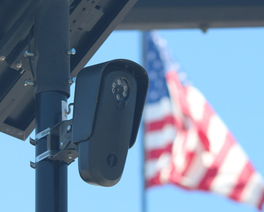 Flock camera monitoring a parking lot as an american flag waves in the background.