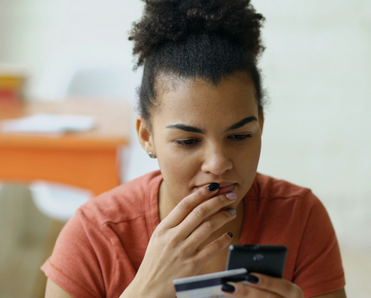 A young woman contemplates her phone with bank card in hand.