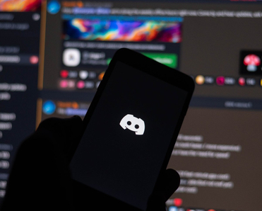 Discord logo on phone screen, with the desktop app opened on a monitor in the background.