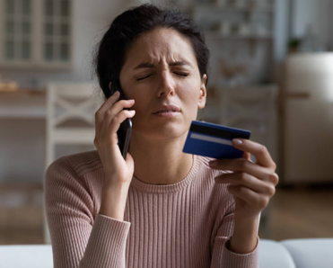 Frustrated woman sitting on couch at home trying to cancel subscription over the phone while holding a credit card