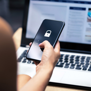 A woman&#x27;s hand holds her cell phone with a locked icon on the screen as she sits in front of her laptop
