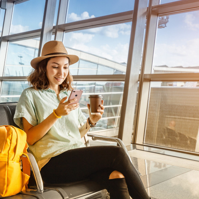 A young Asian woman sits next to her airport gate while using a cell phone and drinking coffee.
