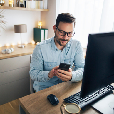 A white man with a beard and glasses sits in his home office and uses his desktop computer and cell phone