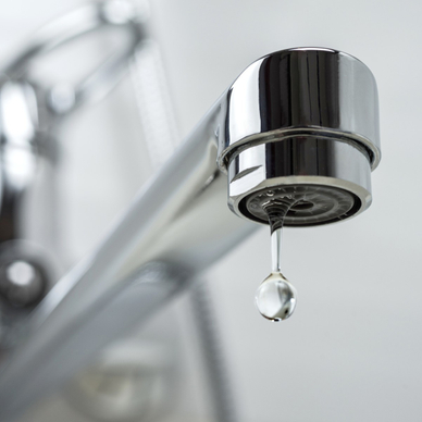 A close-up photo of a stainless steel kitchen sink faucet with a single drop of water leaking out of it.