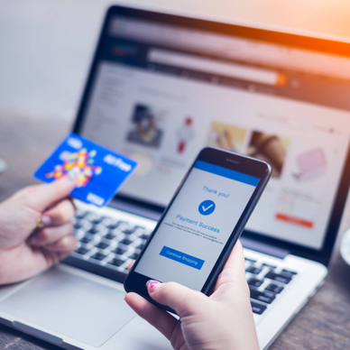 A young woman holds her smartphone that is showing payment success on screen in one hand and her credit card in another, while her laptop is on the table.