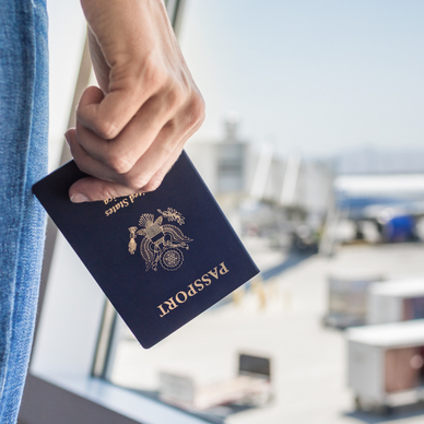 A close-up of a man&#x27;s hand holding a U.S. passport and standing in front of an airport window watching travelers board a white and blue airplane.