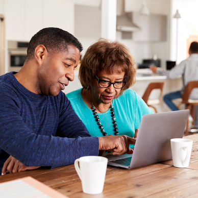 A middle-aged black man helps his older mother use social media safety tips on their home computer.