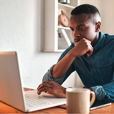 Man thinking, typing, and working on a laptop at his desk with a coffee mug by his side.