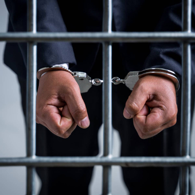 A close-up of a man&#x27;s hands in handcuffs as he&#x27;s behind a jail cell.