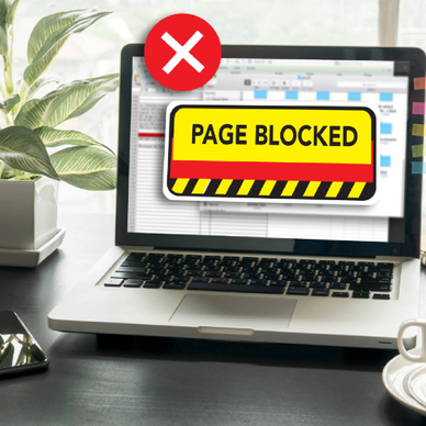 An open laptop on a work desk with the screen displaying the text &quot;page blocked&quot; over a restricted website