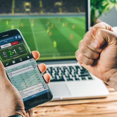 A man holds a phone showing a sports score of 2 to 1 on a betting app. His right fist is clenched in victory. There&#x27;s a laptop screen in the background showing the soccer game being played.