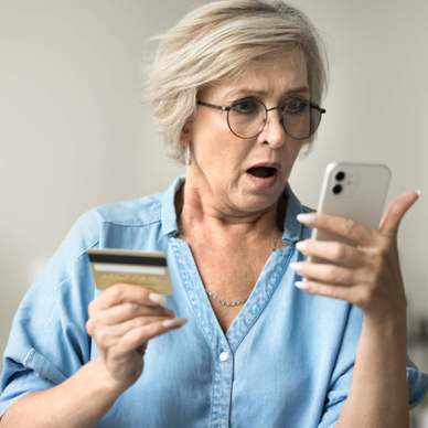 A shocked older woman is holding her smartphone in one hand and her credit card in another.