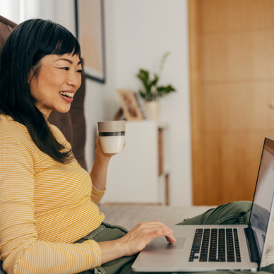 A woman holds a coffee mug and uses her laptop in bed.
