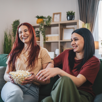 Two women are eating popcorn and laughing on the couch while watching TV.