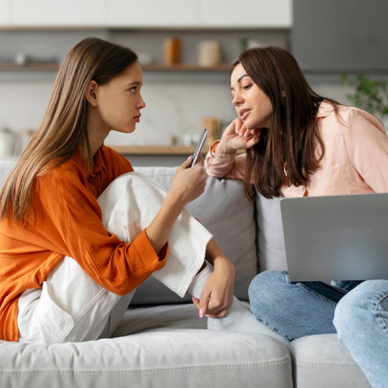 Mother sitting on sofa with laptop spying on teen daughter using cellphone, mom and kid spending leisure time together on devices.