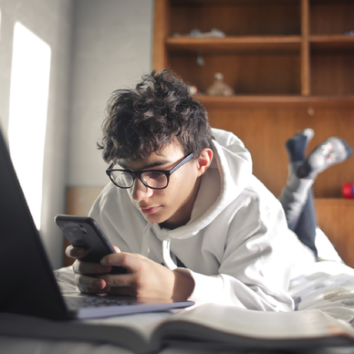 Teen boy laying on bed while scrolling on his smartphone and working on his laptop.