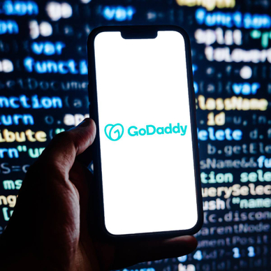 Closeup of smartphone displaying GoDaddy logo with code on a screen in background