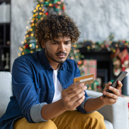 A man is sitting on his couch, holding his phone and his credit card while looking confused.