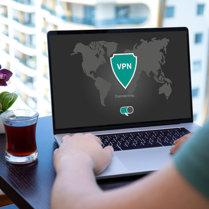 A person types on a laptop computer next to a glass of tea. The laptop screen shows a shield icon that says "VPN." The screen also shows a switch that shows the VPN is turned on.