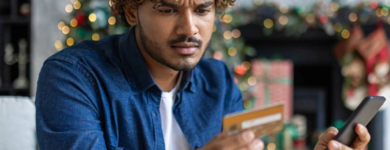 A man is sitting on his couch, holding his phone and his credit card while looking confused.