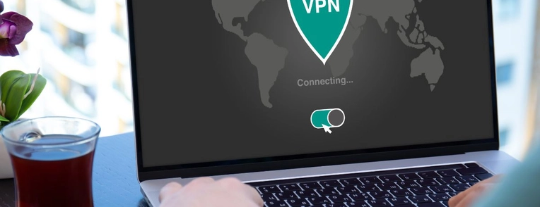 A person types on a laptop computer next to a glass of tea. The laptop screen shows a shield icon that says "VPN." The screen also shows a switch that shows the VPN is turned on.